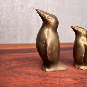 May include: Two antique brass penguin figurines. The penguins are standing upright with their beaks pointed upwards. The brass has a warm, aged patina. The figurines are displayed on a wooden surface.