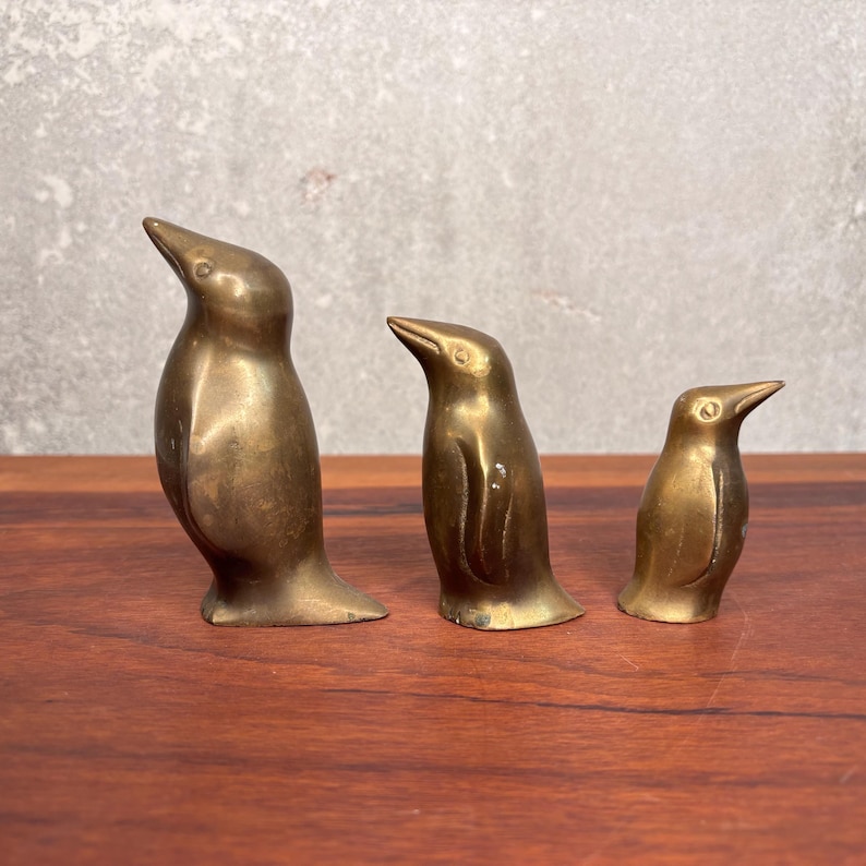 May include: Three brass penguin figurines of varying sizes. The penguins are standing upright with their beaks pointed upwards. The figurines have a shiny, metallic finish and are arranged on a wooden surface.