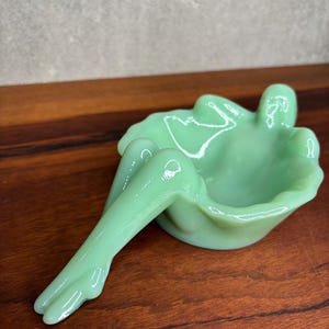 May include: A light green ashtray shaped like a reclining figure. The ashtray features a shallow bowl and an extended leg. It is made of a glossy, opaque material. The ashtray is displayed on a wooden surface.