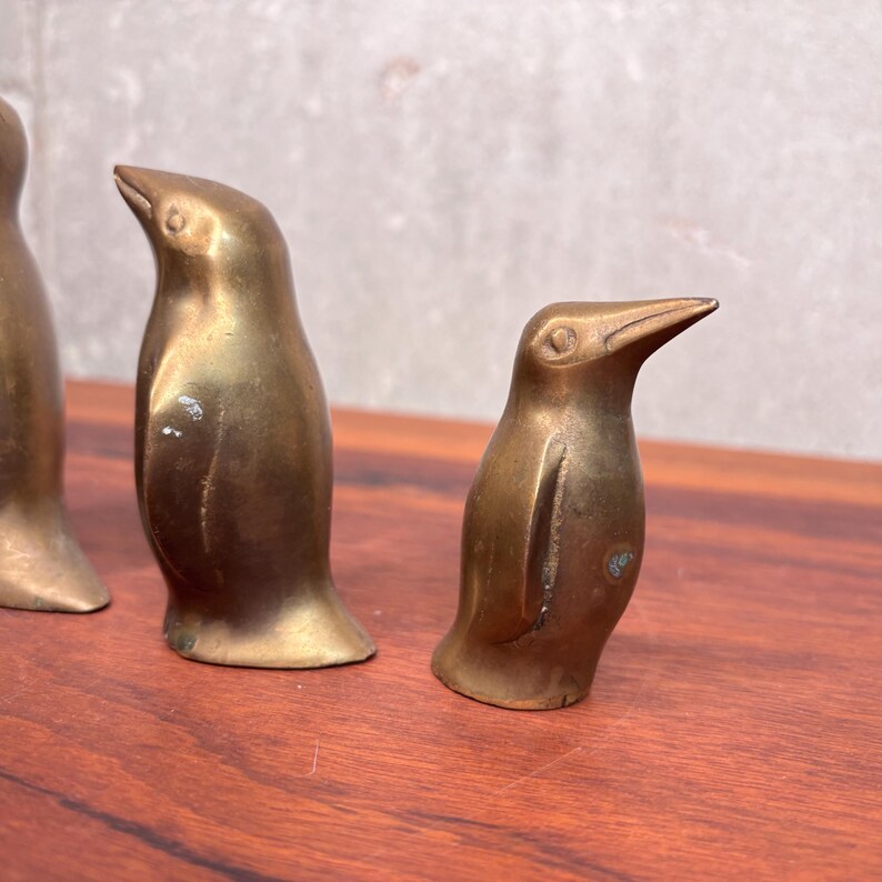 May include: Three vintage brass penguin figurines. The penguins are in various sizes and poses, with a warm, golden hue. They are displayed on a wooden surface, adding a touch of retro charm to any decor.