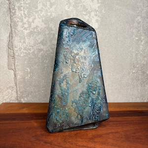 Puede incluir: Un jarrón de cerámica alto y trapezoidal con un acabado texturizado multicolor. La superficie presenta una mezcla de tonos azul profundo, bronce metálico y carbón oscuro con un esmalte rugoso e irregular que crea un aspecto complejo.