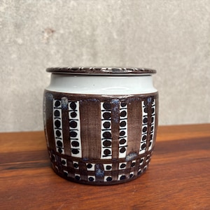 May include: A ceramic jar with a lid featuring a white upper band and a brown lower section decorated with vertical stripes of black circles. The base of the jar displays a grid pattern of black squares and circles on a dark brown background.
