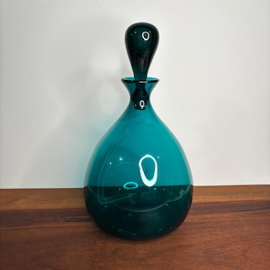 May include: A teal glass bottle with a teardrop shape and a matching stopper. The bottle has a dark teal base and a lighter teal upper section. The stopper is a dark teal color.