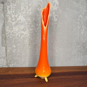 May include: A vibrant orange glass vase with a distinctive flared top and a rounded base. The vase features a long, slender neck and rests on three small, cream-coloured feet. The interior of the vase is a lighter shade of orange.