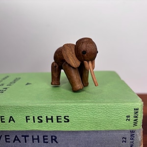May include: A small, wooden elephant figurine with a trunk and large ears. The elephant is brown and stands on four legs. It is placed on top of a stack of green and gray books. The books have text on the spines.