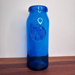 May include: A cobalt blue glass bottle with a round, embossed design. The bottle has a narrow neck and a wide base. The embossed design appears to be a stylized bird or emblem. The bottle is sitting on a wooden surface.