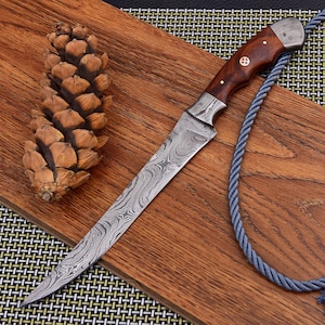 Könnte beinhalten: Ein Damaststahlmesser mit Holzgriff und dekorativer Zwinge liegt auf einem Holzschneidebrett. Das Messer hat eine lange, schlanke Klinge mit einem einzigartigen Muster. Ein Tannenzapfen und ein blaues Seil befinden sich ebenfalls auf dem Brett.