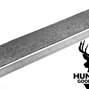 Può includere: Una lama di metallo rettangolare a motivi con un righello per la scala. La lama ha un design unico e strutturato. Il righello mostra le misure in centimetri. Il logo Hunting Good Store si trova in basso a destra.