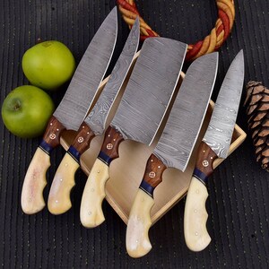 Puede incluir: Un juego de cinco cuchillos de cocina de acero de Damasco. Las hojas presentan un diseño distintivo. Los mangos son de madera con empuñaduras color hueso, detalles azules y dorados. Los cuchillos están colocados en una bandeja de madera.