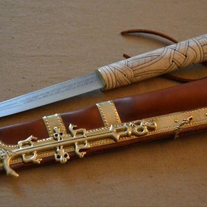 Op de afbeelding: Een vaststaand mes met een zilverkleurig stalen lemmet en een crèmekleurig handvat met gedetailleerde zwarte lijntekeningen. Het mes wordt geleverd met een bruine lederen schede versierd met sierlijke goudkleurige metalen beslag en klinknagels.