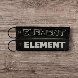 Könnte beinhalten: Zwei schwarze Schlüsselanhänger mit silbernen Metallringen. Die Schlüsselanhänger sind aus Stoff und haben den Schriftzug "ELEMENT" in weiß aufgedruckt.