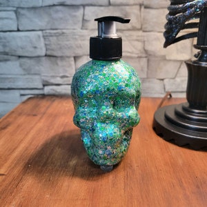 May include: A skull-shaped soap dispenser with a black pump top. The dispenser is covered in green, blue, and silver glitter. The product is sitting on a wooden surface.