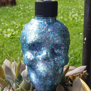 May include: A blue glitter skull-shaped soap dispenser with a black pump top. The dispenser is decorated with iridescent glitter and sits atop a bed of green and purple succulents. The dispenser is a unique bathroom accessory.