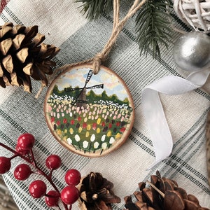 May include: A hand-painted wooden ornament featuring a windmill and a field of colorful tulips. The ornament is attached to a twine string and is perfect for adding a touch of whimsy to your holiday decor.