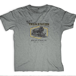 May include: A gray t-shirt featuring a vintage train graphic within a yellow hexagonal frame. The text reads "Rip's Train Station Tours" and "Montana Wyoming Line One Way Ticket". The design is in black and yellow.