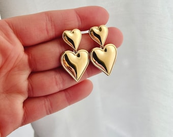May include: Gold tone earrings with three heart-shaped charms. The hearts are stacked on top of each other, with the largest heart at the bottom.