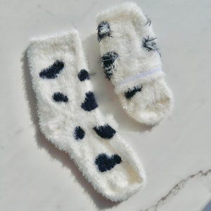 May include: A pair of white fuzzy socks with black spots. The socks are made of a soft, fluffy material and have a comfortable fit.
