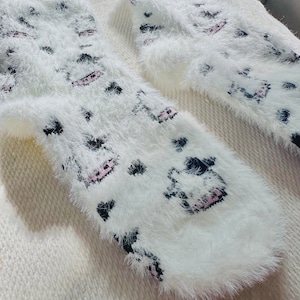May include: A pair of white fuzzy socks with a black and pink cartoon design. The socks are soft and fluffy.