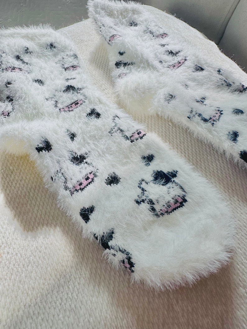 May include: A pair of white fuzzy socks with a black and pink cartoon design. The socks are soft and fluffy.