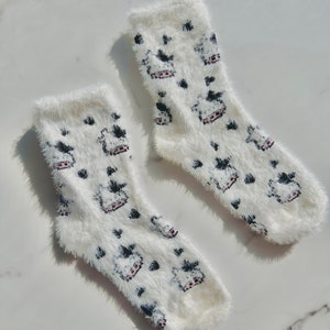 May include: A pair of white fuzzy socks with a black and pink cow print design.
