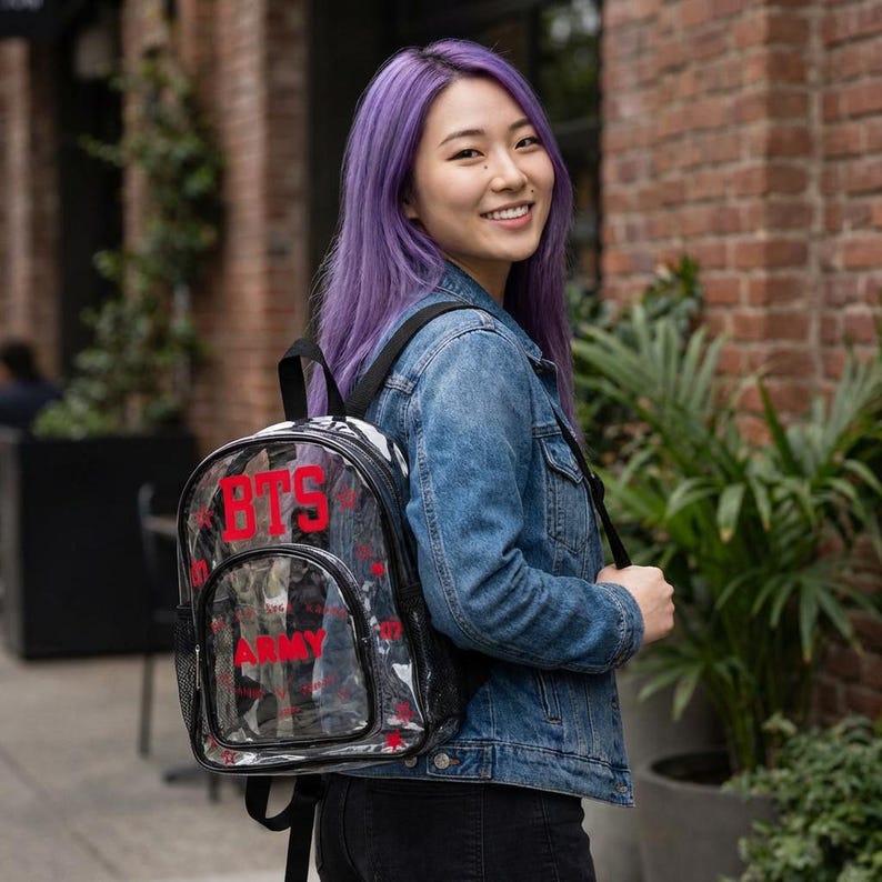 K&ouml;nnte beinhalten: Ein durchsichtiger Rucksack mit dem roten Text "BTS ARMY" wird von einer Person getragen. Der Rucksack hat schwarze Verzierungen und eine Seitentasche aus Netzstoff. Die Person tr&auml;gt eine Jeansjacke und eine schwarze Hose. Der Hintergrund umfasst Ziegel und Gr&uuml;n.