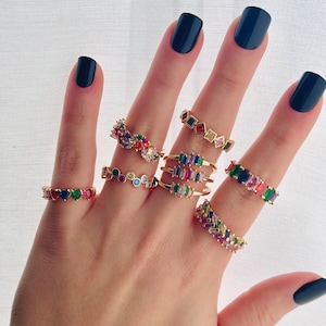 May include: A hand wearing multiple colorful gemstone rings. The rings are made of gold and feature different shapes and sizes of gemstones. The hand is wearing black nail polish.