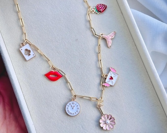 May include: A gold chain necklace with multiple charms, including a strawberry, a mermaid tail, a playing card, a red lip, a letter with a heart, a clock face, and a pink flower.