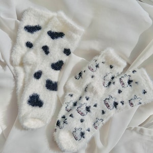 May include: Two pairs of fuzzy white socks with black and white patterns. One pair has a cow print and the other has black spots.