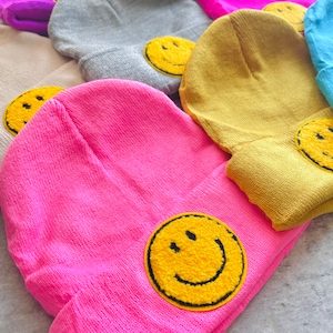May include: A collection of colorful knit beanies with a yellow smiley face embroidered on each one. The beanies are arranged in a pile, with the pink beanie on top.