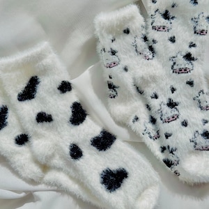 May include: Two pairs of fuzzy white socks with black and white cow print designs. One pair has small black hearts and the other has small cow silhouettes.