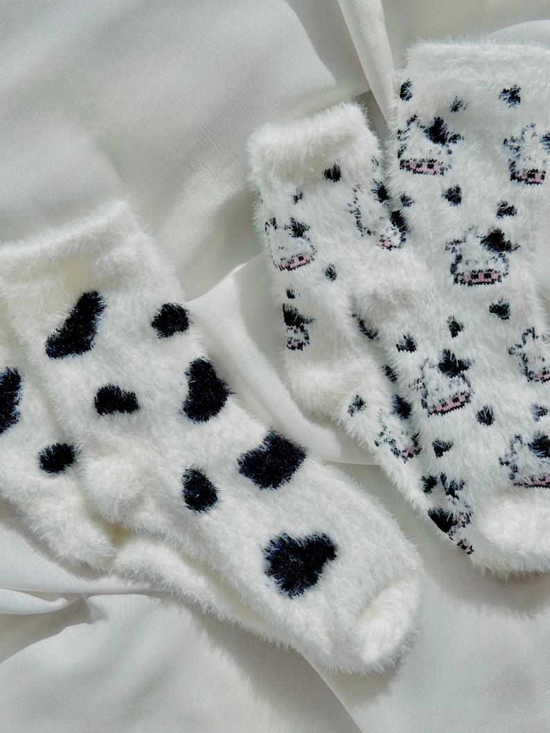 May include: Two pairs of fuzzy white socks with black and white cow print designs. One pair has small black hearts and the other has small cow silhouettes.