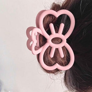 Puede incluir: Una pinza para el pelo rosa claro con un diseño en forma de corazón. La pinza está hecha de plástico y presenta una serie de formas de corazón y un diseño central. La pinza sujeta una porción de cabello castaño.