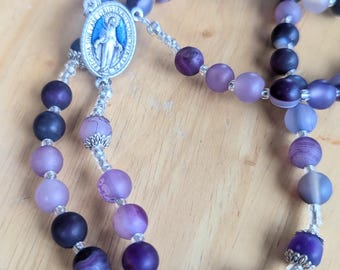 May include: A purple and black rosary with a silver centrepiece featuring a depiction of the Virgin Mary. The beads are made of agate and have a variety of shades of purple.