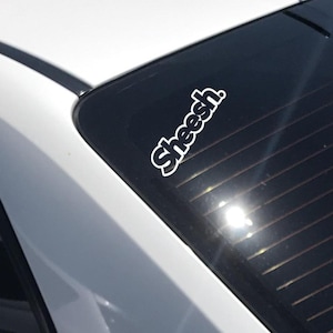 Puede incluir: Coche blanco con ventana trasera negra. Dos pegatinas de vinilo blanco están adheridas a la ventana. La pegatina superior dice "Sheesh". La pegatina inferior es un oso de dibujos animados con estrellas en los ojos.