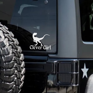 Può includere: Un adesivo in vinile bianco applicato sul lunotto posteriore di un SUV nero. L'adesivo presenta la sagoma di un dinosauro velociraptor posizionata sopra il testo "Clever Girl" scritto in un carattere serif. Il veicolo ha una ruota di scorta.