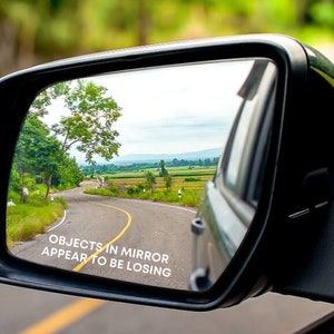 May include: A car's wing mirror showing a country road with fields and trees under a cloudy sky. The text "OBJECTS IN MIRROR APPEAR TO BE LOSING" is displayed on the mirror.