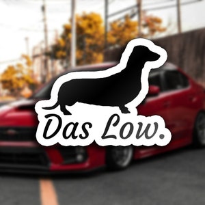 Puede incluir: Una pegatina blanca con una silueta negra de un dachshund y el texto "Das Low." La pegatina está colocada en un coche rojo.