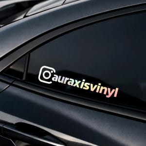May include: A holographic vinyl decal applied to a dark car window, featuring the Instagram camera icon followed by the text auraxisvinyl in a lowercase sans-serif font with a shifting iridescent color gradient across the letters.