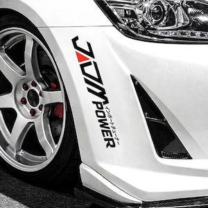 May include: A white car featuring a black and red "JDM POWER" sticker on the front bumper. The sticker includes Japanese text. The car has a silver wheel with red detailing.