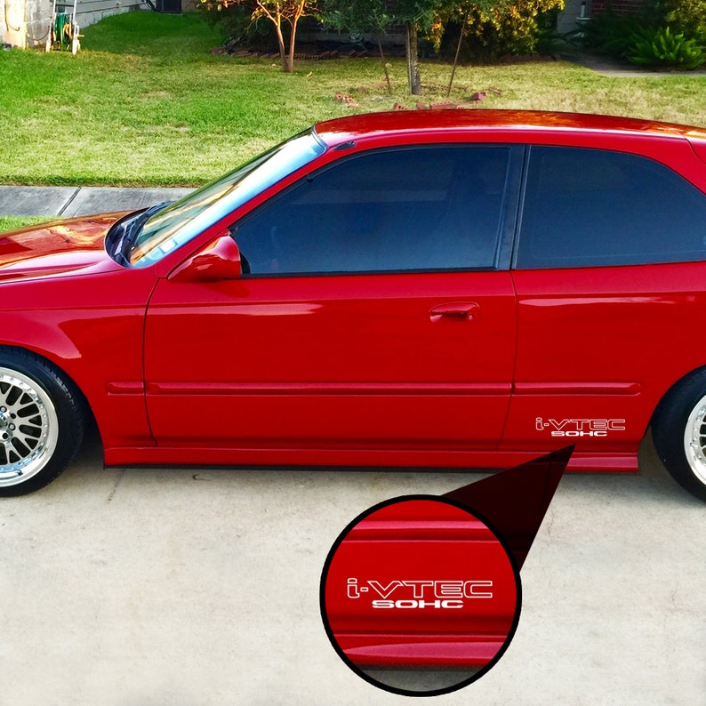 May include: A red car with tinted windows and silver rims. The side of the car features white text that reads "i-VTEC SOHC". The car is parked on a concrete surface with grass in the background.