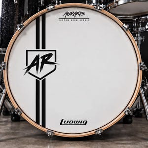 Könnte beinhalten: Ein weißes Bassdrum-Fell mit einer schwarzen Grafik, die die Buchstaben AR in einem Schild zeigt, flankiert von zwei vertikalen schwarzen Streifen. Der Text Auraxis Custom Drum Decals steht oben und das Ludwig-Logo befindet sich unten.