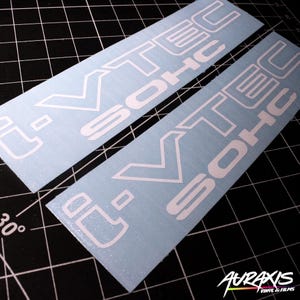 May include: Two white vinyl decals with the text "i-VTEC SOHC" in a bold, stylized font. The decals are rectangular and have a light blue background. The image is taken on a black grid surface.