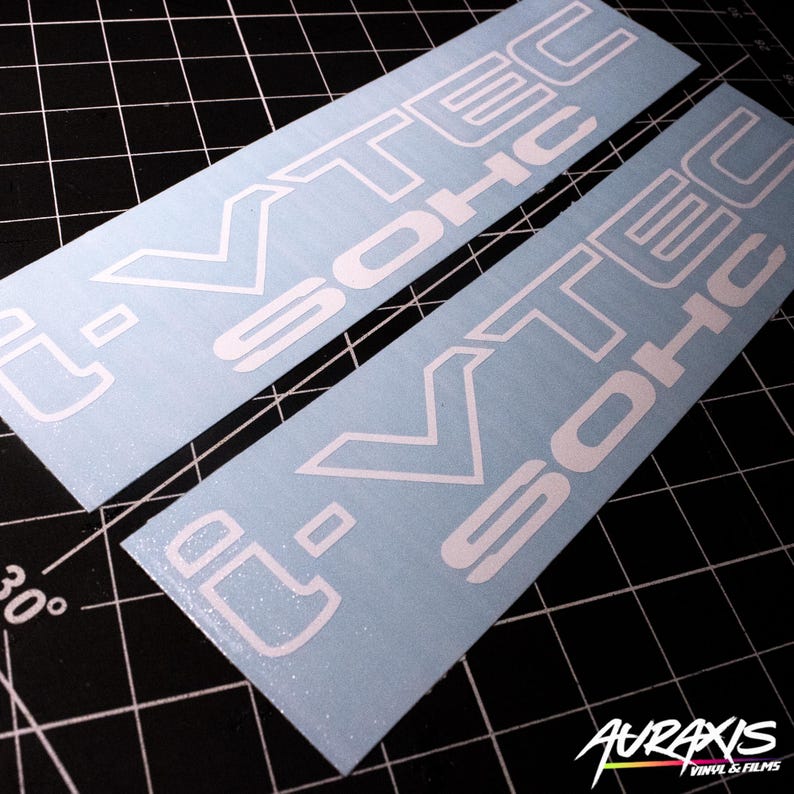 May include: Two white vinyl decals with the text "i-VTEC SOHC" in a bold, stylized font. The decals are rectangular and have a light blue background. The image is taken on a black grid surface.
