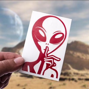 Op de afbeelding: Een rood alien-ontwerp op een witte rechthoekige sticker. De alien heeft grote ovale ogen en een vinger op zijn lippen in een 'shush'-gebaar. De achtergrond toont een woestijnlandschap en een planeet.