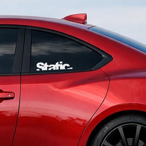 May include: White "Static" decal on the rear window of a red car. The decal is in a bold, sans-serif font. The car has a black trim around the window and a black tire.