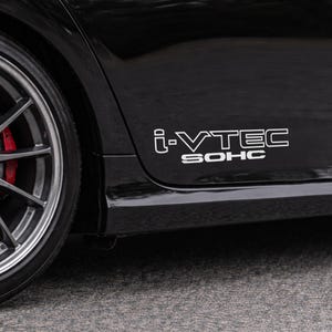 May include: Close-up of a black car with a silver wheel and red brake caliper. The car's side panel displays the white text "i-VTEC SOHC". The image is taken outdoors on a gray surface.