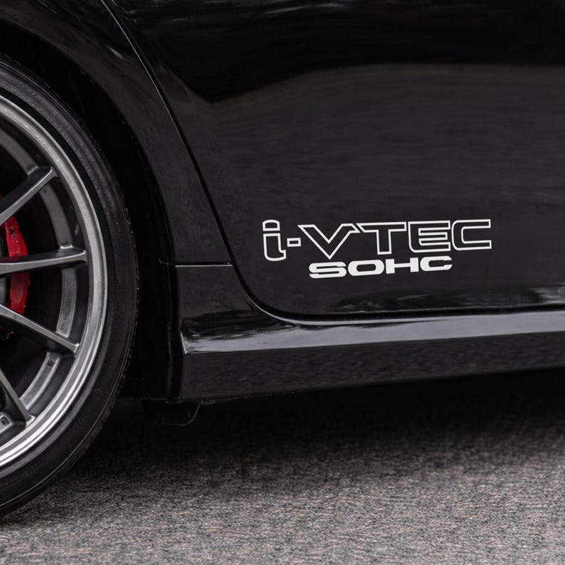 May include: Close-up of a black car with a silver wheel and red brake caliper. The car's side panel displays the white text "i-VTEC SOHC". The image is taken outdoors on a gray surface.