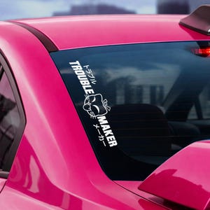 May include: A white vinyl decal applied to the rear window of a bright pink car. The decal features a stylized cat face graphic positioned between the English text TROUBLE MAKER and the corresponding Japanese katakana characters for trouble maker.