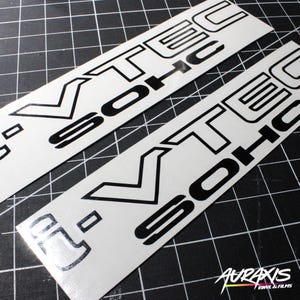 May include: Two white vinyl stickers with the text "VTEC SOHC" in black. The stickers are rectangular and have a glossy finish. The background is a black grid.
