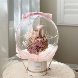 May include: A clear balloon filled with dried flowers and greenery, tied with a pink ribbon. The balloon is attached to a white box with the text "Ballons & Co." printed on it.